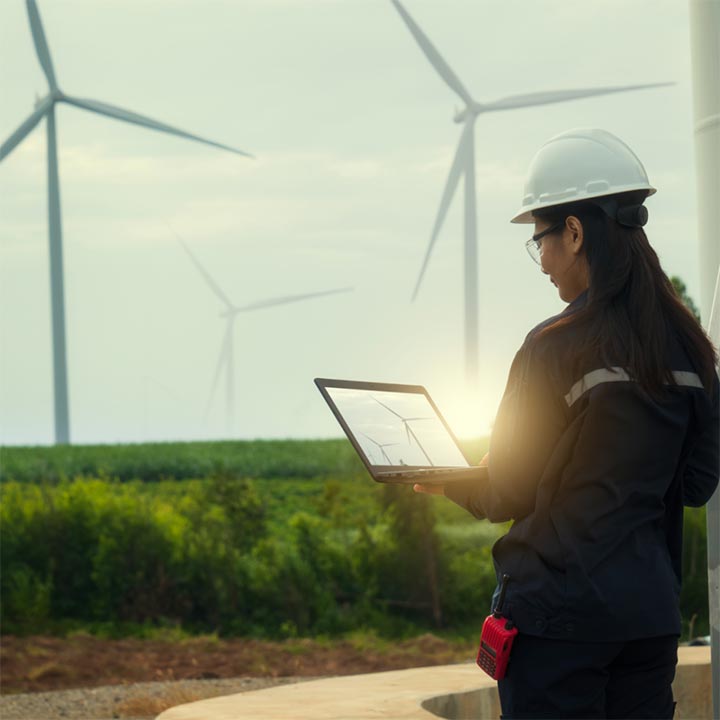 Woman wearing safety helmet and glasses, looking at a laptop in front of field of wind turbines