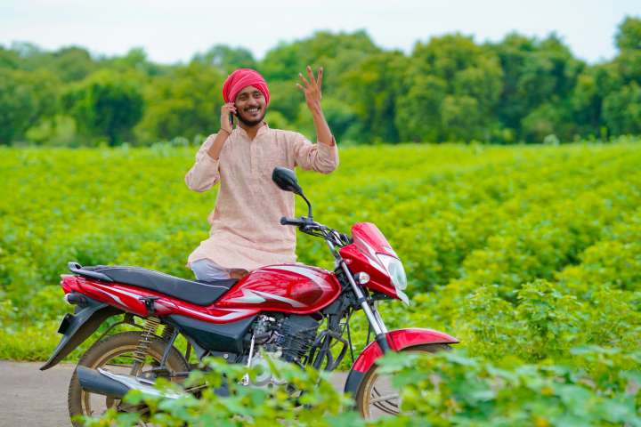 An Indian man on his phone, standing by a motorbike by the side of a field